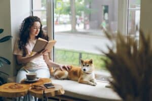 vrouw zit in het raam en leest een boek met een hond naast haar. op tafel voor haar staat een kop koffie en ligt een telefoon